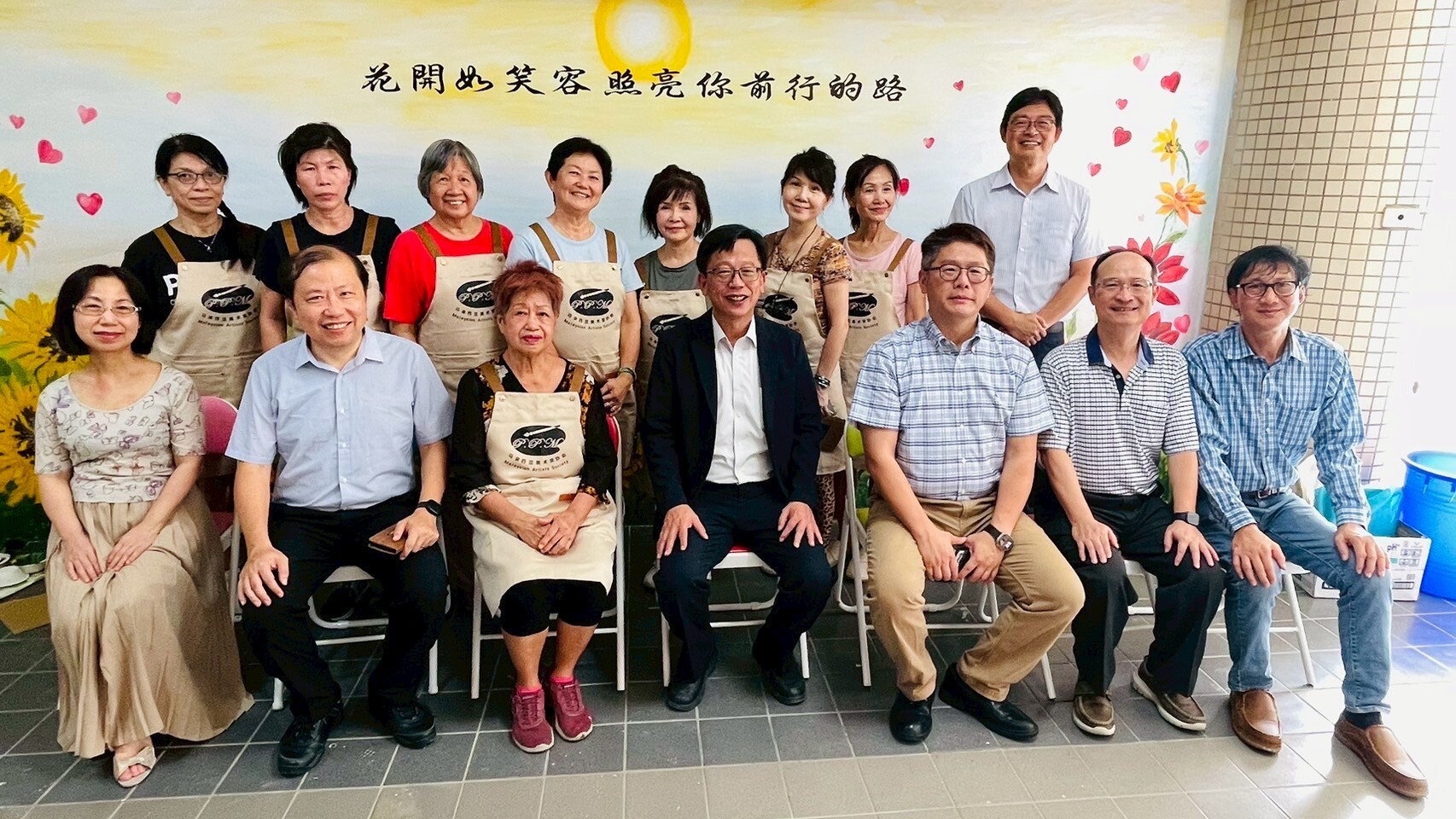 本校師長與馬來西亞藝術家們在另一幅壁畫前合影，畫作上寫著：「花開如笑容，照亮你前行的路」。NCUE faculty members posed for a group photo with the Malaysian artists in front of another mural, which bears the inscription: “Flowers bloom like smiles, lighting the path ahead of you.” 